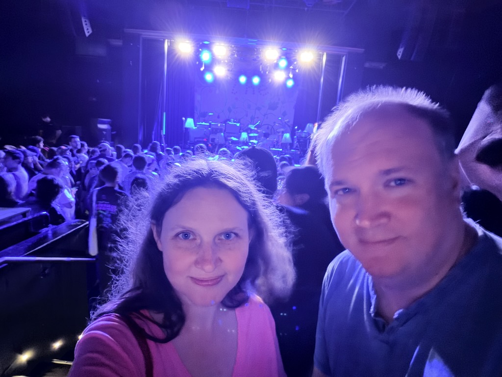 Shannon and Jerrod at an indoor concert venue. It is dark and the stage is behind us. The concert hasn't started yet.